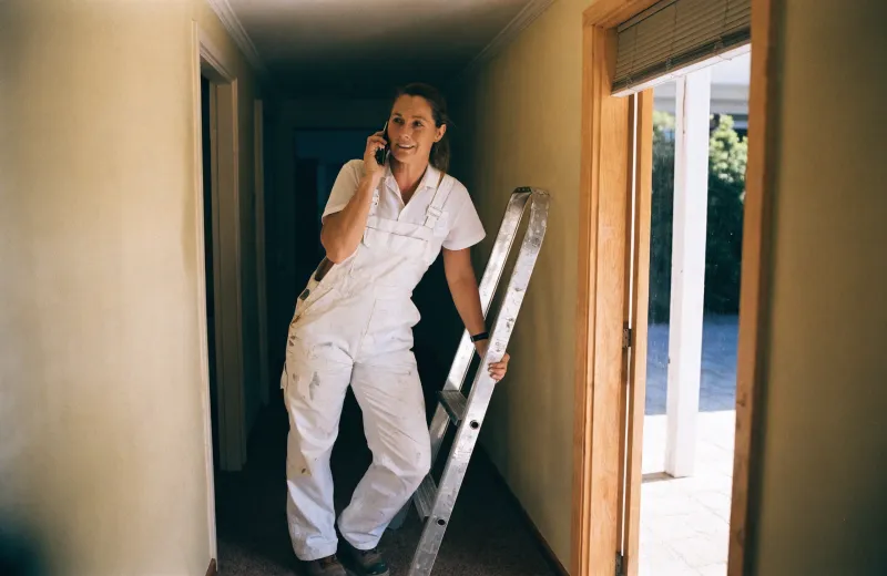 Australian painter in overalls taking a phone call while moving a ladder in a hallway